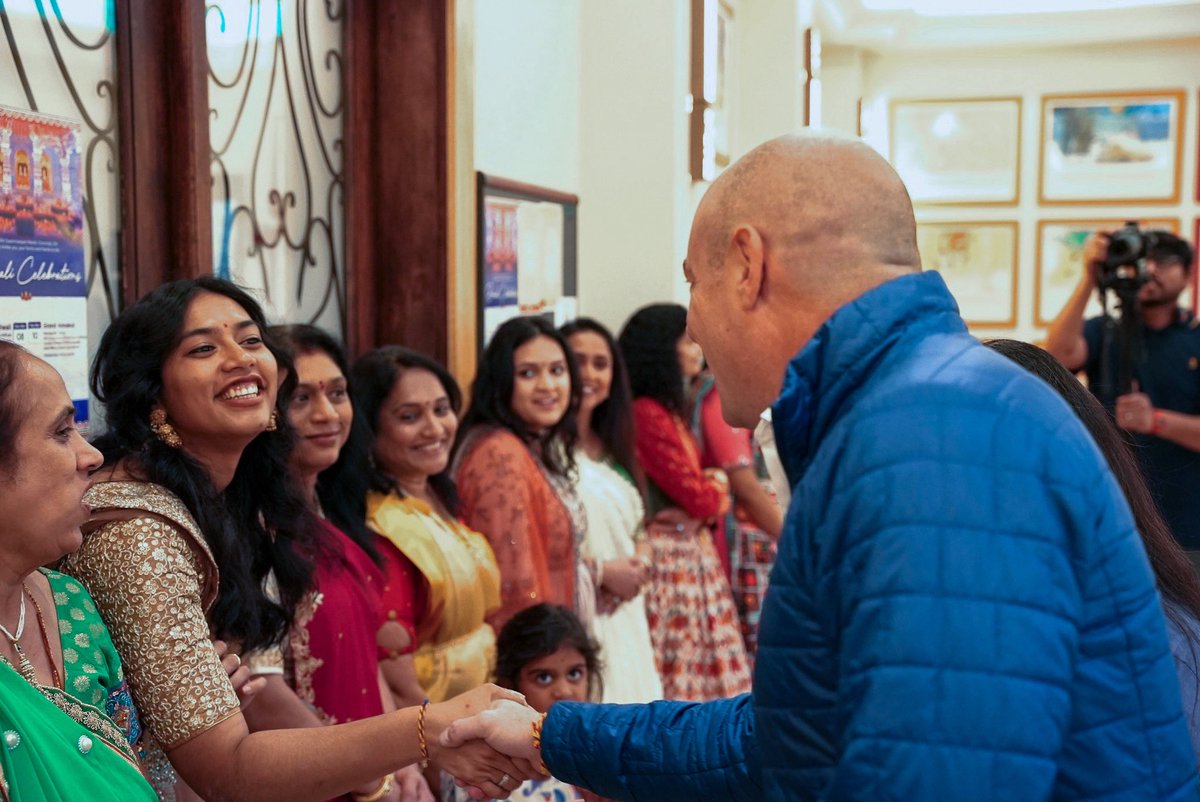 A bald man in a blue jacket greets and shakes hands with a woman in a line of women at an indoor event. A photographer is taking pictures in the background, and posters and framed pictures are visible on the walls.
