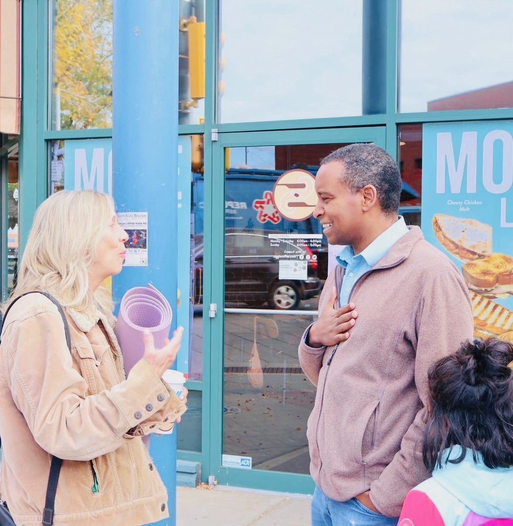Two adults talk outside a storefront while a child is partially visible in the foreground. The woman holds a rolled purple item and a drink, and the man smiles with one hand on his chest. Storefront signs and other text are visible.