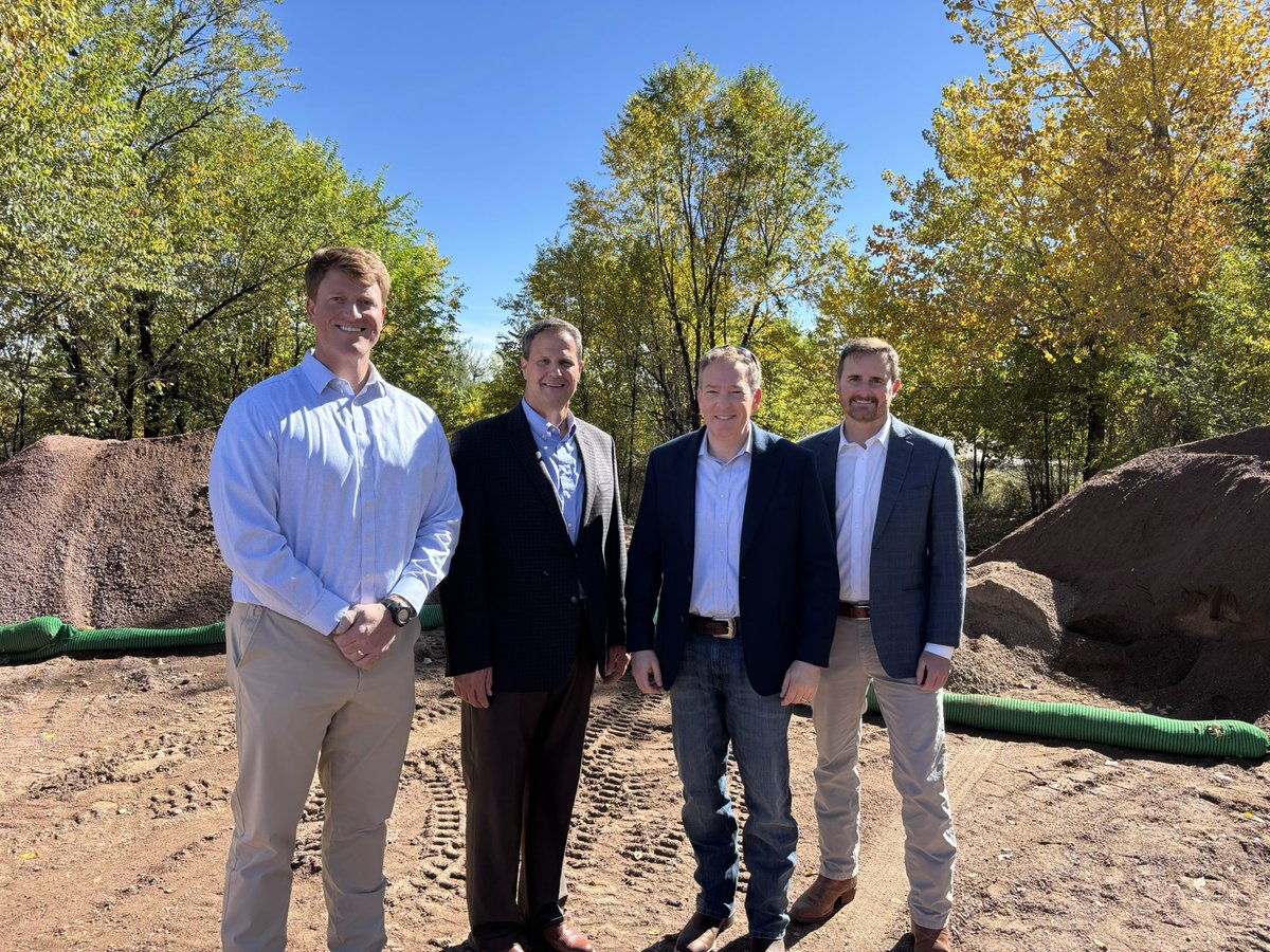 Four men in business attire pose together at an outdoor construction or landscaping site. Trees with autumn foliage and piles of soil are behind them. No text is visible.