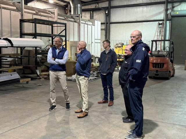 Five men stand inside an industrial workshop or warehouse, looking toward something off camera. Machinery, shelving, materials, and a forklift are visible in the background. There is some text on clothing and equipment.