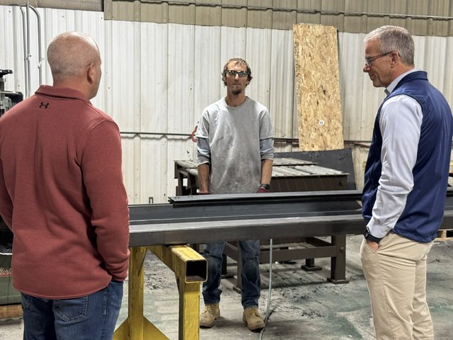 Three men stand around a long metal beam in a workshop or industrial building. One man is behind the beam, while the other two face him. There is small text on clothing and equipment.