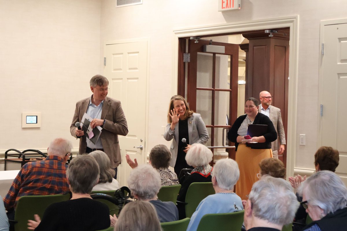 A group of people stand at the front of a room while an audience seated in rows of chairs applauds. One woman waves, and two people hold microphones. An exit sign is visible above a doorway.