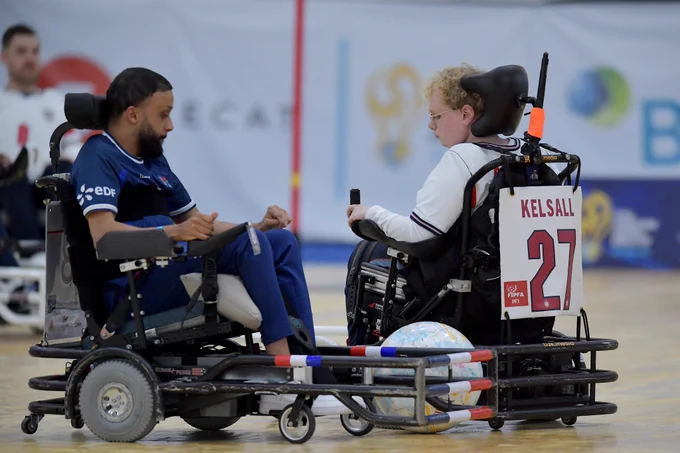 Two powerchair football players compete for the ball during an international match on an indoor court. France player Bryan Weiss, wearing a blue kit, faces England player Kelsall, whose name and number 27 are displayed on the back of the powerchair. The ball sits between the two chairs as they contest possession.