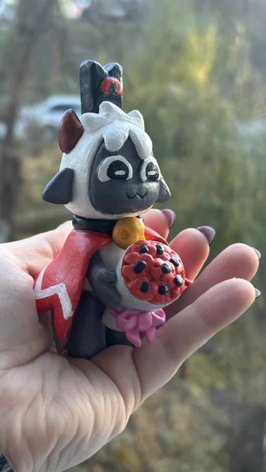 A handmade polymer clay figurine of the Lamb from Cult of the Lamb, held in a hand against a blurry nature background. Showcased from side views.