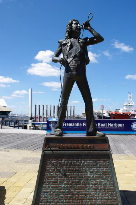 Bronze statue of Bon Scott singing into a raised microphone, standing on a bronze Marshall amp atop an inscribed plinth, with the Fremantle Fishing Boat Harbour sign, boats and blue sky behind