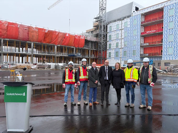 Representatives from the Government of Saskatchewan and Graham Construction Limited stand together at the Regina Specialized Long-Term Care Home project site. The group poses in front of the multi-storey building under construction, with scaffolding, a crane and construction materials visible in the background.