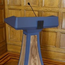 Identifies as Podium. Maybe the most expensive non-custom-made lectern on the market. About 80% sass. Hobbies: Sitting in a corner collecting dust.