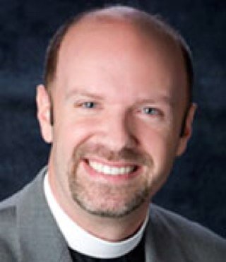 Seen wearing a bow tie, he is pastor at Holy Trinity Lutheran Church. When making a video for the church, makeup had to be used to reduce the shine on his head.