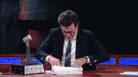 Stephen Colbert sitting at a desk frantically flipping throu